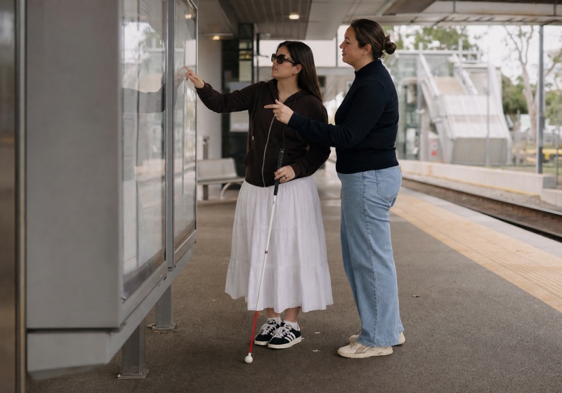 Two women reading a timetable together at a train station, one holding a white cane