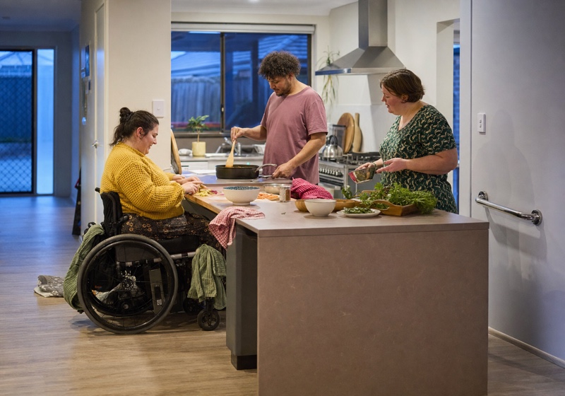 Three people preparing a meal together in a shared kitchen, one of them using a wheelchair